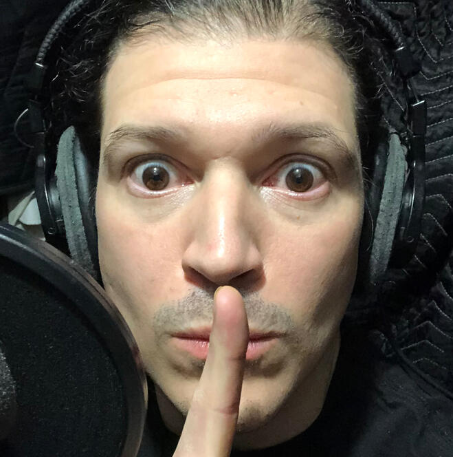 A wide-eyed pale man holds a finger to his lips as if to shush you.